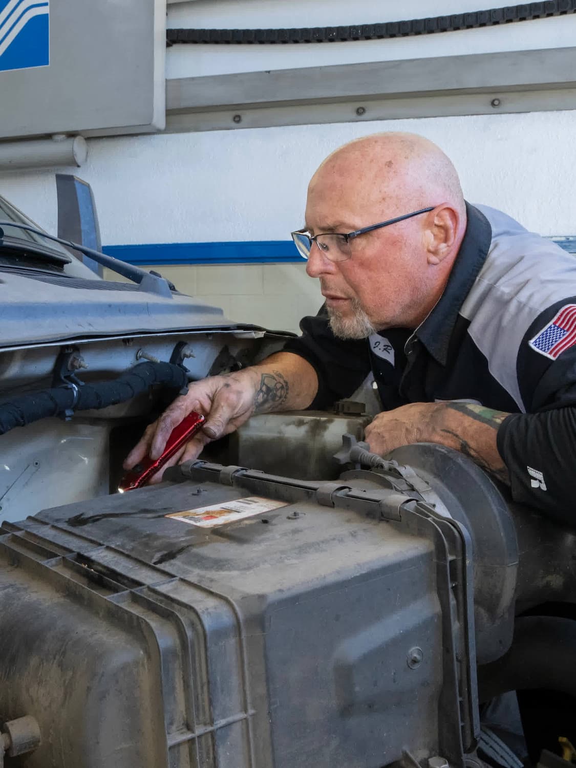 A mechanic with glasses and a bald head inspects an engine, holding a red tool. He wears a work uniform with an American flag patch on the sleeve.