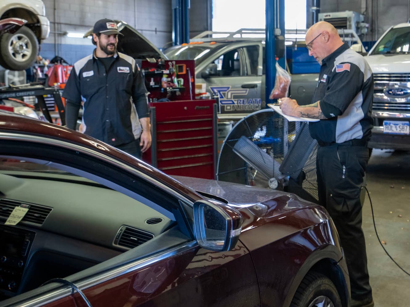 Two auto mechanics in a garage, one writing on a clipboard and the other standing nearby, surrounded by vehicles and tools. An open car door is visible in the foreground.