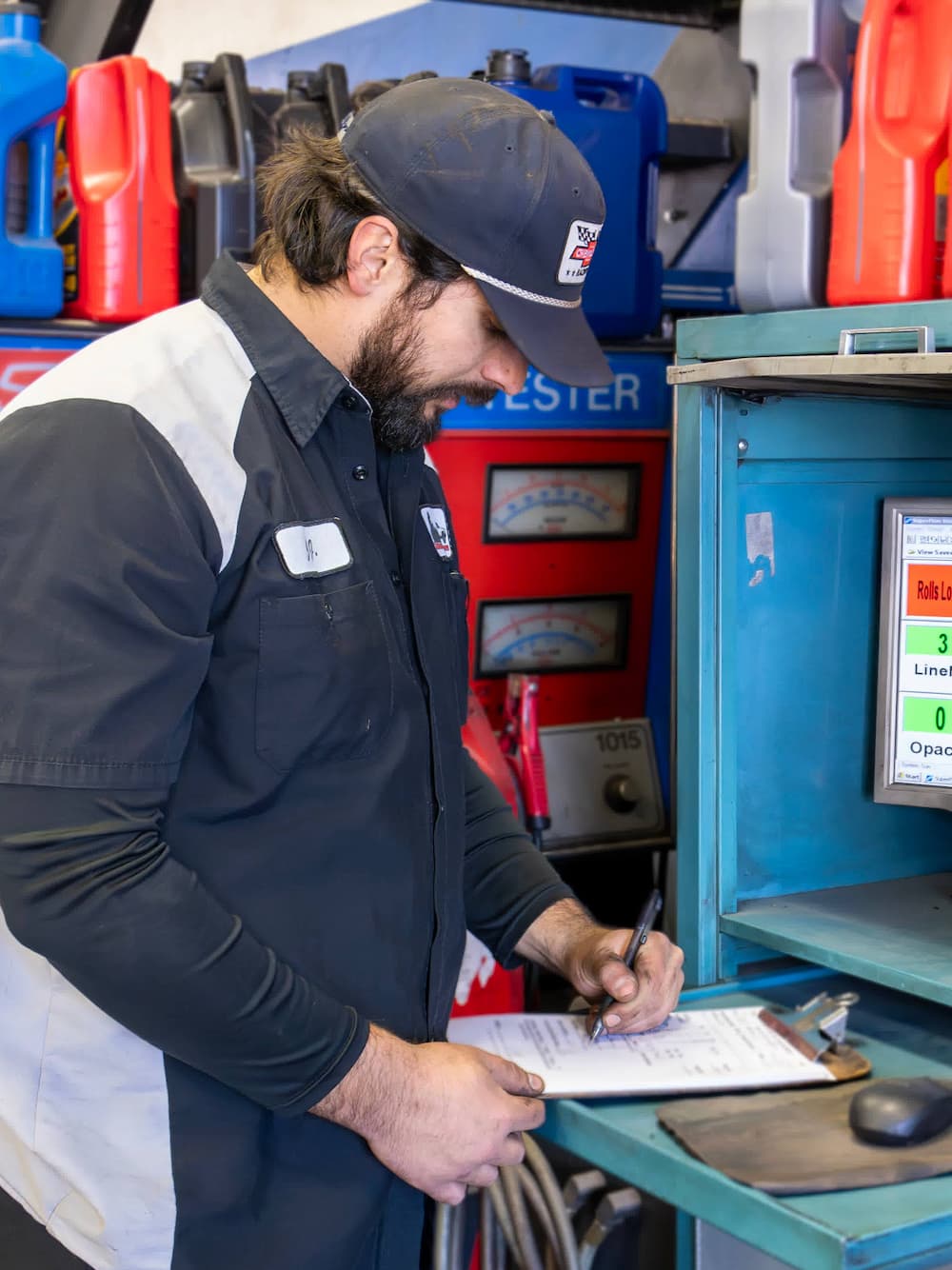 A mechanic wearing a cap and uniform writes on a clipboard in an auto shop, surrounded by tools, containers, and equipment. A monitor and gauges are visible in the background.