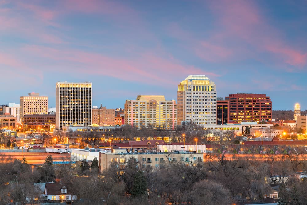 Downtown city skyline at sunset with modern buildings, warm lights illuminating the buildings, and a partly cloudy sky with pink and blue hues in the background. Trees and smaller buildings are visible in the foreground.