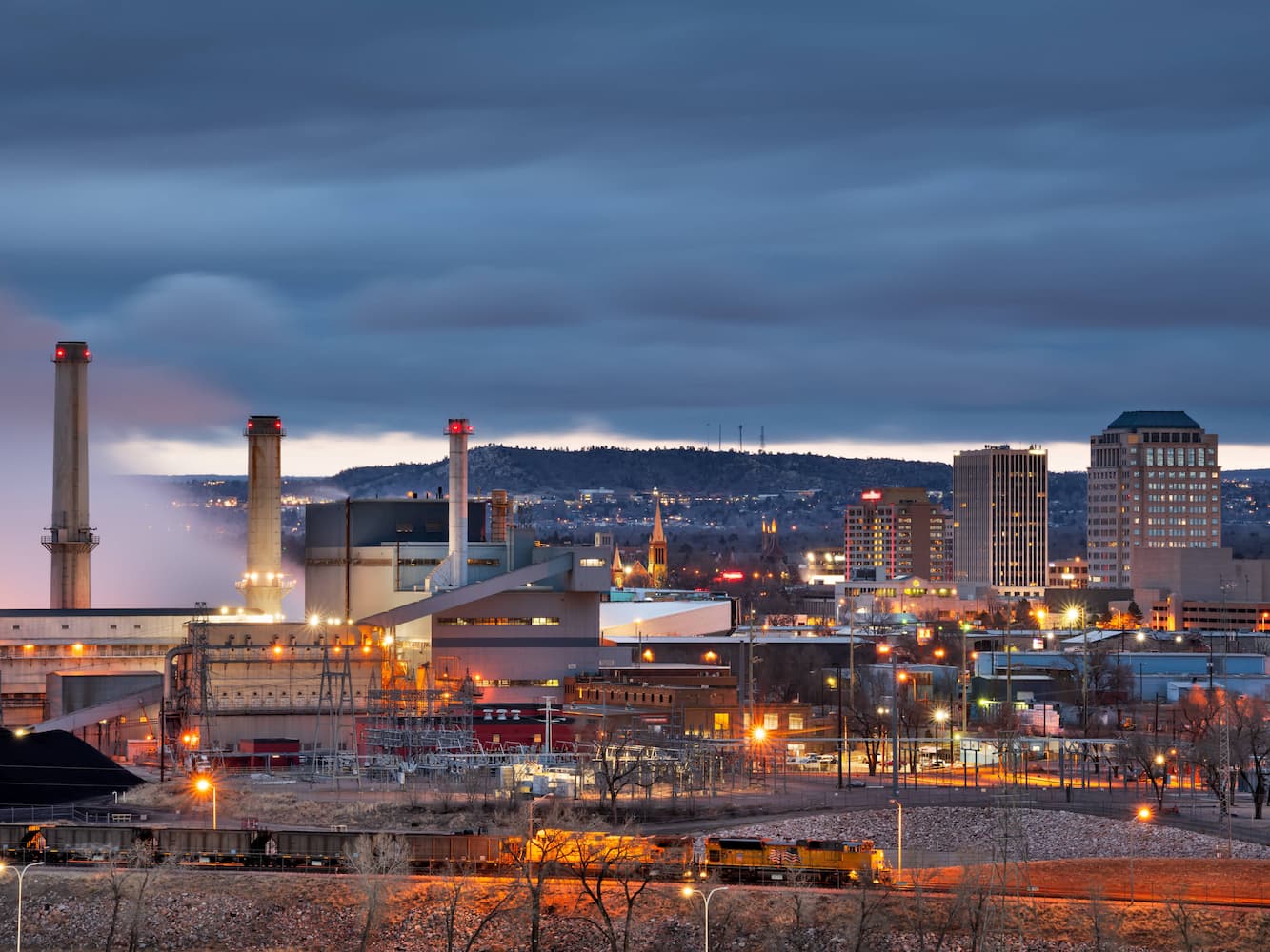 Cityscape at dusk showing an industrial area with factories and smokestacks in the foreground, and illuminated buildings and a cloudy sky in the background.